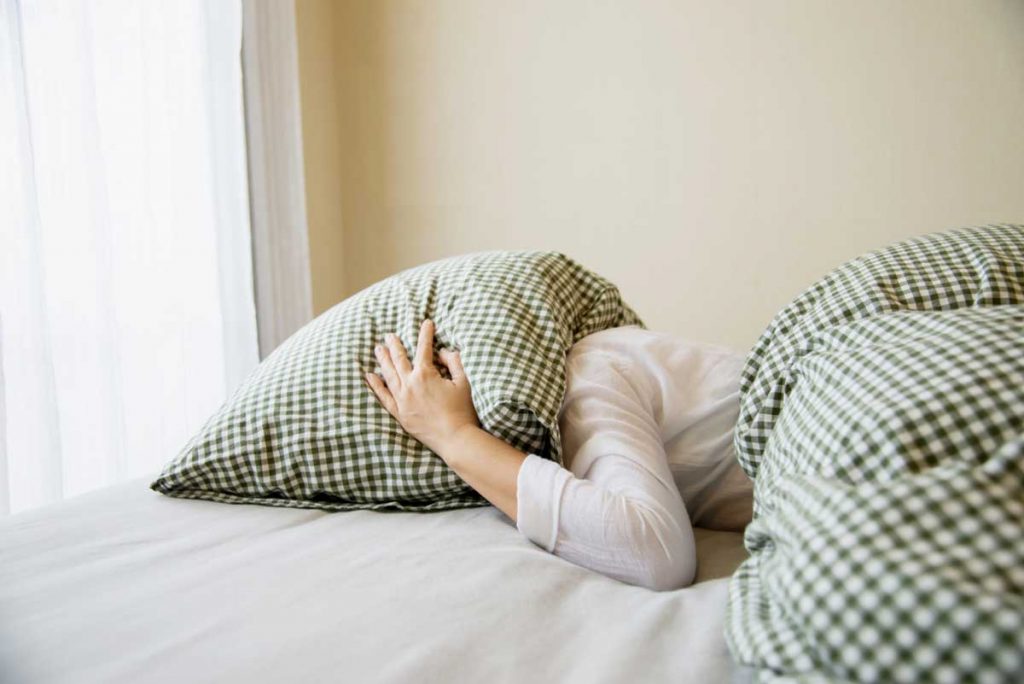 kobieta cierpiąca na nadwrażliwość słuchową leży w łóżku z poduszką nałożoną na głowę