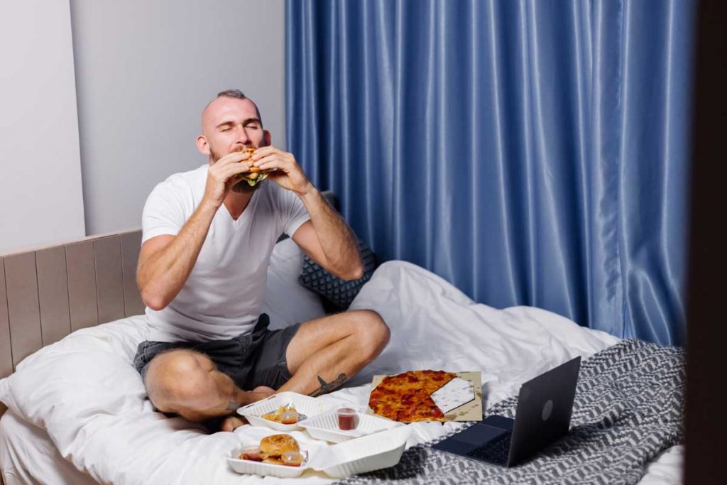 młody mężczyzna siedzący na łóżku otoczony niezdrowym jedzeniem typu fast food
