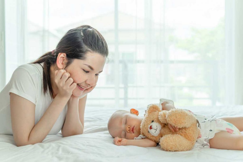 uśmiechnięta mama patrząca z czułością na swoje śpiące na materacu niemowlę